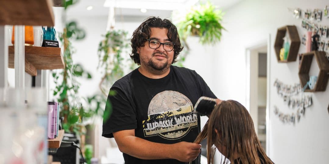 Image of a man working in his hair salon, brushing a woman’s hair and smiling at the camera.