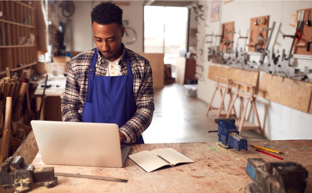 Man wearing a blue apron working on a laptop in a woodworking workshop, with tools and equipment in the background