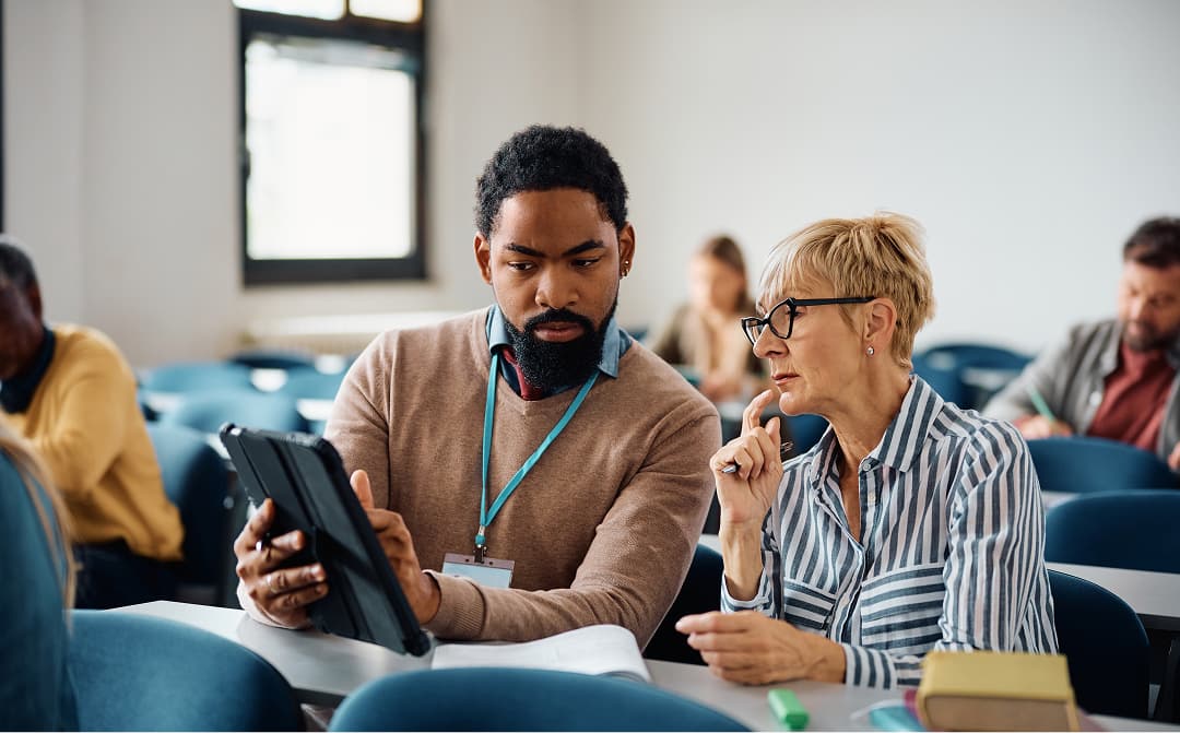 Young man with a tablet explaining something to an older woman in a classroom
